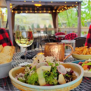 a restaurant patio table laden with pottery bowls filled with salad, bread, and pasta. diners are enjoying white wine and a mule with cozy buffalo plaid blankets