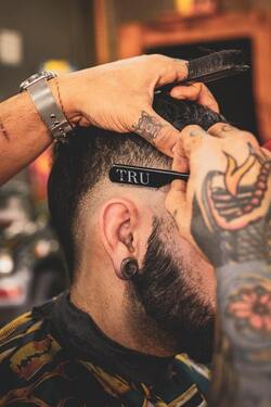 A barber with tattooed hands finishing a fade on a bearded client